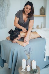 Eine Masseurin gibt einer Kundin auf einer Massagebank eine entspannende Rückenmassage in einem stilvoll eingerichteten Raum mit Kerzen und Lavendel-Dekoration.